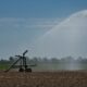 Large agricultural sprinkler irrigating a wide, dry field under a clear blue sky, with water spraying in a high arc across the farmland.