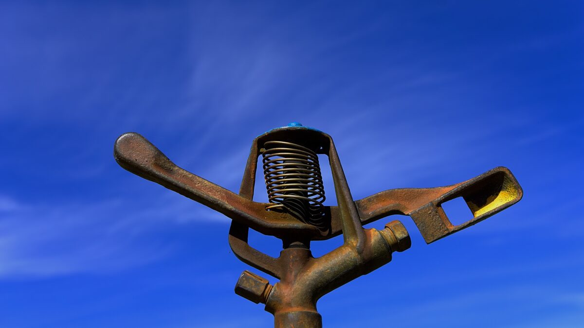 "Close-up of a rusted metal sprinkler head against a clear blue sky, used in agricultural or lawn irrigation systems.