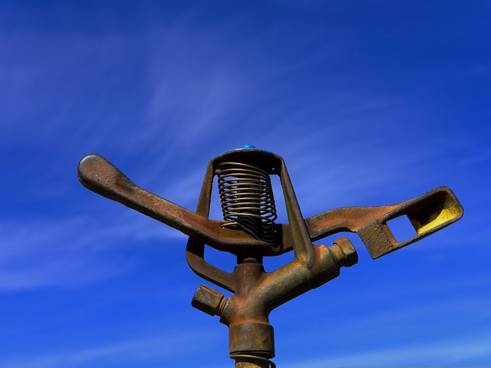 "Close-up of a rusted metal sprinkler head against a clear blue sky, used in agricultural or lawn irrigation systems.